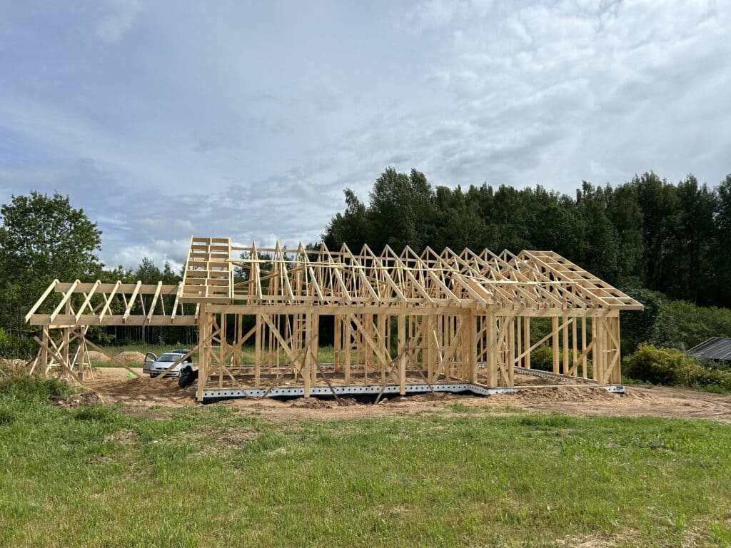 Daļēji uzbūvēts koka mājas karkass stāv uz zāliena zemes gabala, kas uz koku un mākoņainu debesu fona rāda spāres un jumta kopnes. Pie konstrukcijas ir redzama celtniecības tehnika.