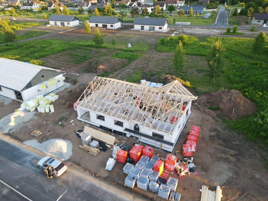 Aeroskats uz daļēji uzbūvētu māju ar koka kopnēm ar redzamu koka karkasu dzīvojamā rajonā. Ap māju atrodas būvmateriālu kaudzes, tai skaitā siltinājums un ķieģeļi. Blakus ir citas mājas un atvērti zemes gabali. Uz ielas novietots transportlīdzeklis.