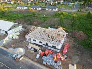 Aeroskats uz daļēji uzbūvētu māju ar koka kopnēm ar redzamu koka karkasu dzīvojamā rajonā. Ap māju atrodas būvmateriālu kaudzes, tai skaitā siltinājums un ķieģeļi. Blakus ir citas mājas un atvērti zemes gabali. Uz ielas novietots transportlīdzeklis.