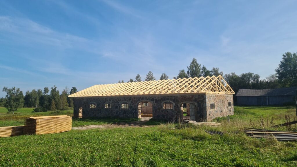 Zālainā laukā zem skaidrām zilām debesīm stāv puscelta mūra ēka ar nepabeigtu koka kopnes jumta karkasu. Netālu atrodas vairākas koka dēļu kaudzes. Fonā redzami koki un cita koka konstrukcija.